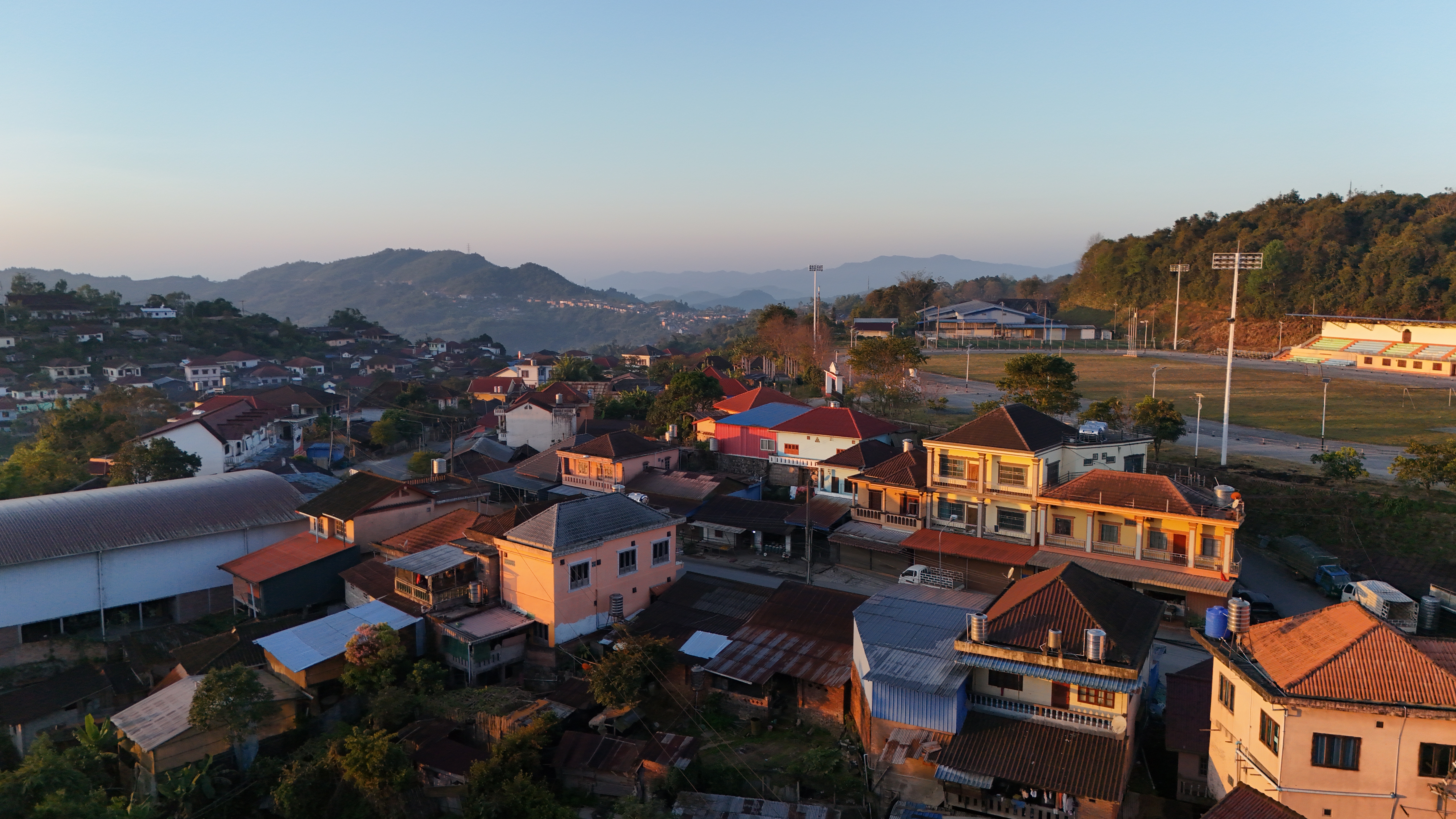 Phongsali at dawn from the drone — red-tile roofs cascading down the ridge in the golden hour.