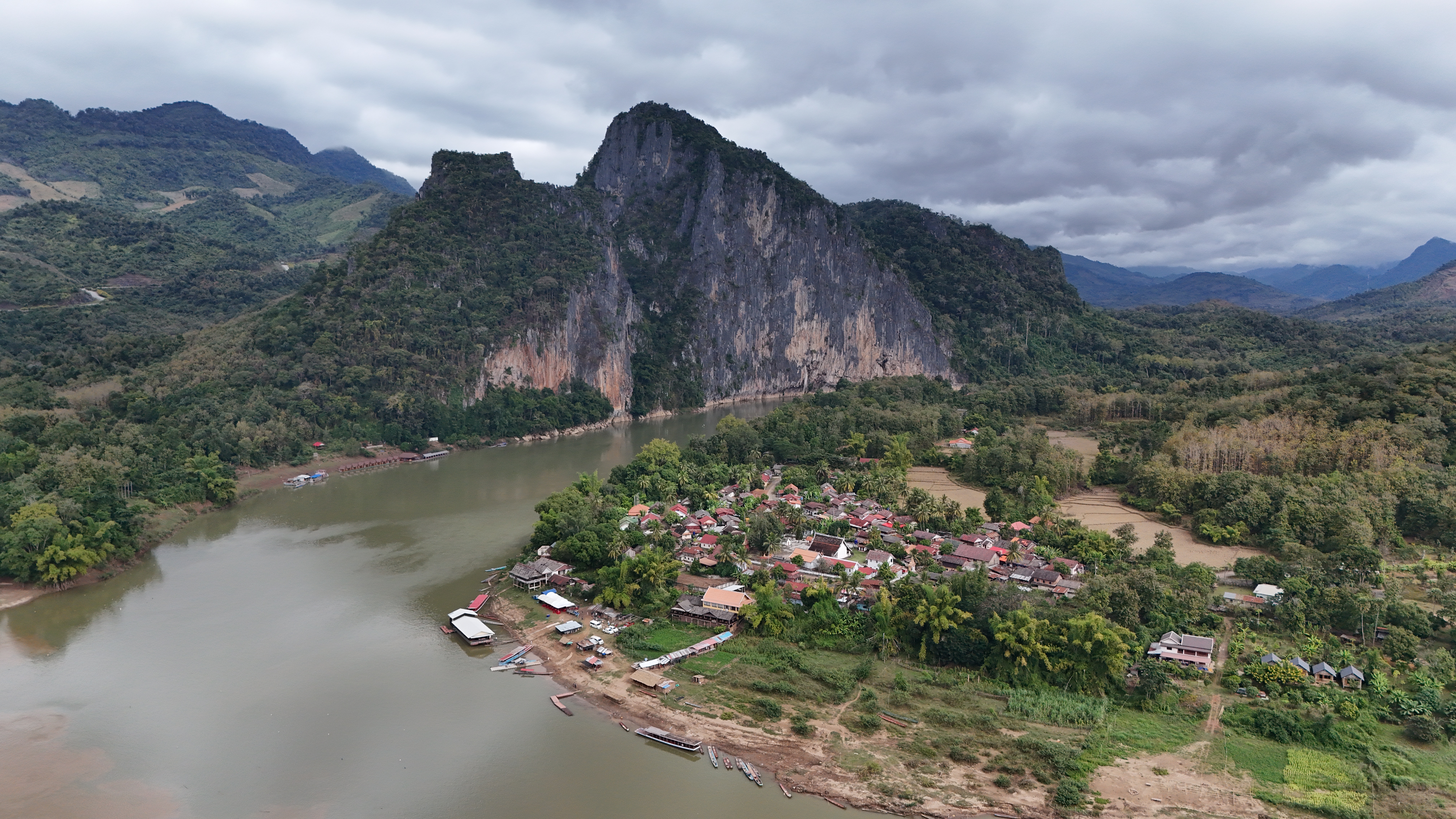 Wider view from the same flight — the Mekong bending past the Pak Ou cliffs, longtail boats lined up at the landing below.